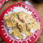 A red plate with a white decorative border holds a serving of white rice topped with a creamy, yellowish sauce and chunks of chicken. The plate is on a wooden table next to a bowl of herbs, salt shaker, and a bowl of rice.