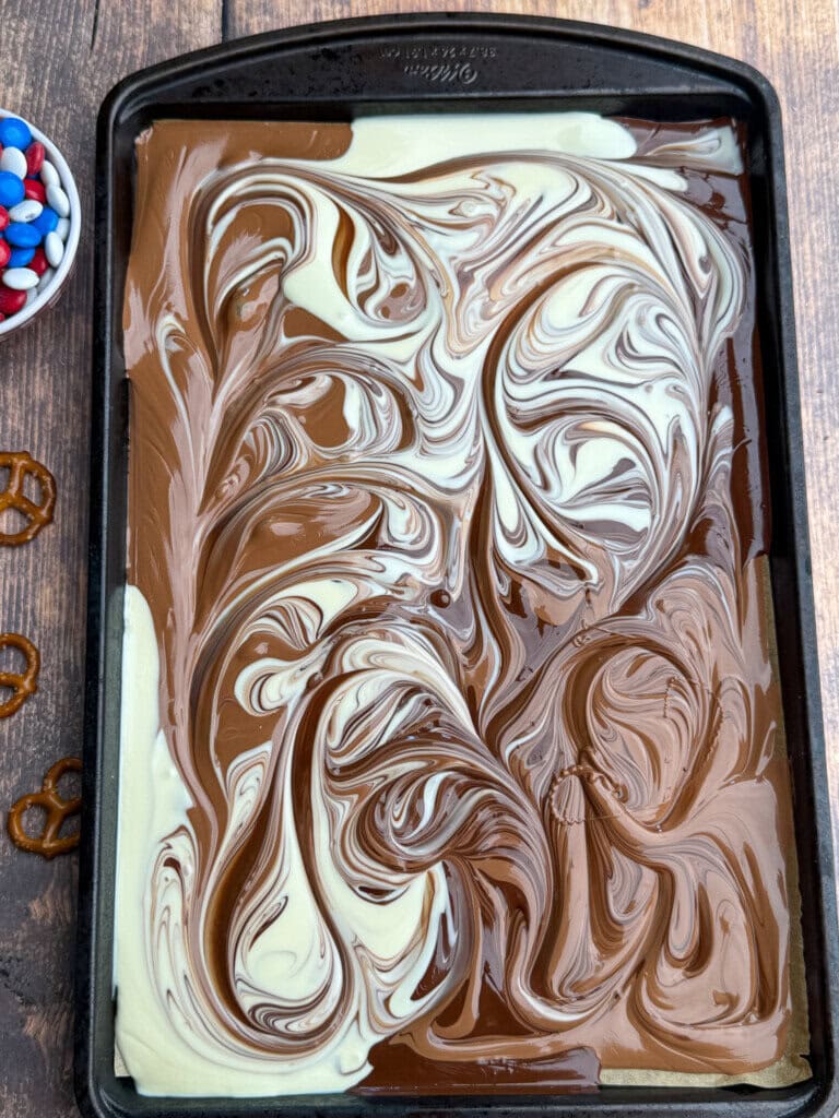 A baking sheet with a marbled mixture of swirled milk and white chocolate. Nearby are red, white, and blue candies in a bowl and a few pretzels on a wooden surface.