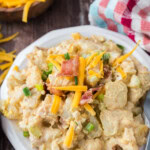 A white plate of creamy potato salad topped with shredded cheddar cheese, green onions, and crispy bacon bits, with a colorful checkered napkin and extra cheese in the background.