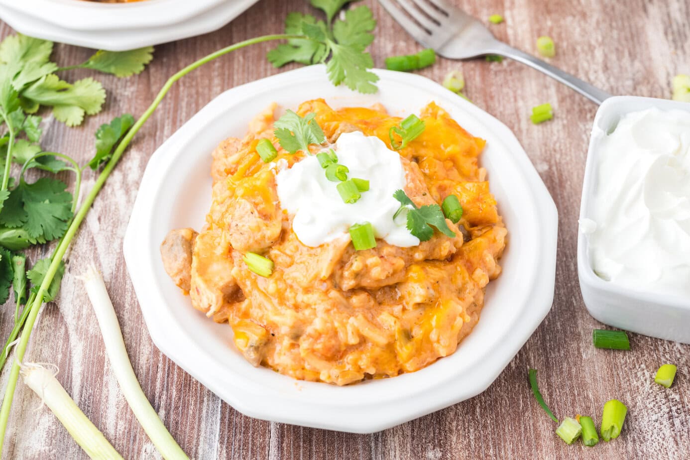 A plate of cheesy chicken and rice casserole topped with sour cream, chopped green onions, and cilantro, served on a white dish with fresh herbs and a fork nearby on a wooden table.