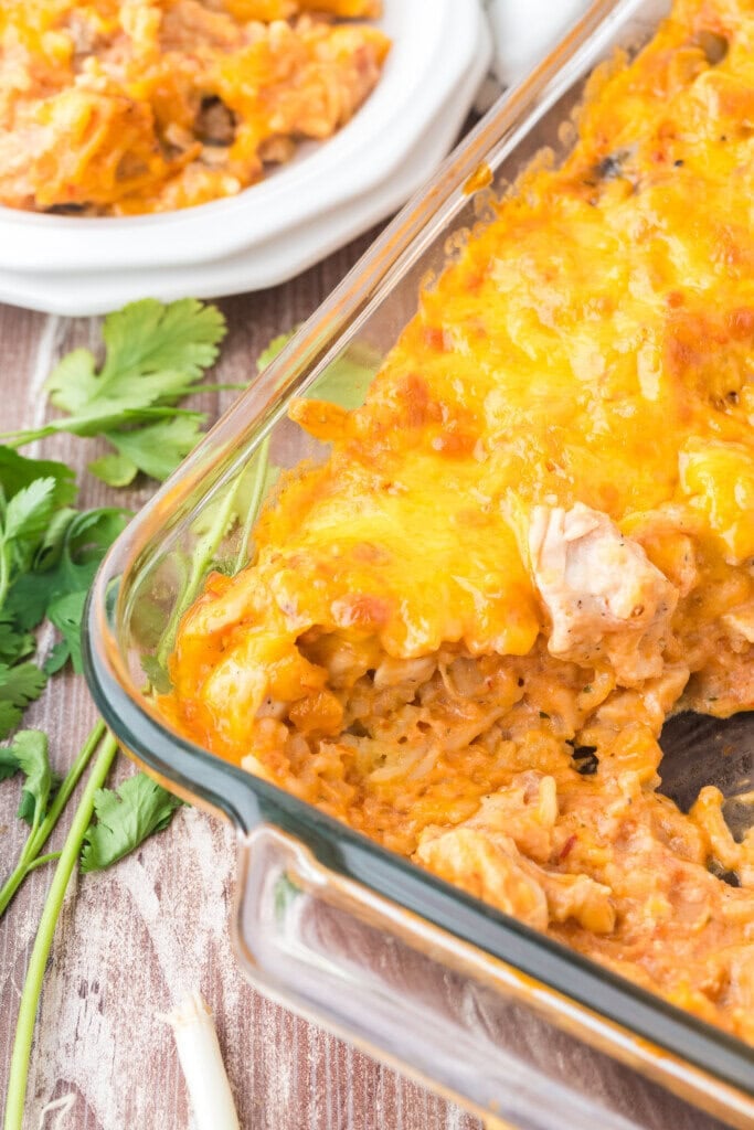 A glass baking dish filled with cheesy chicken and rice casserole, partially served, sits on a wooden table next to fresh cilantro and a plate with a serving of the casserole.