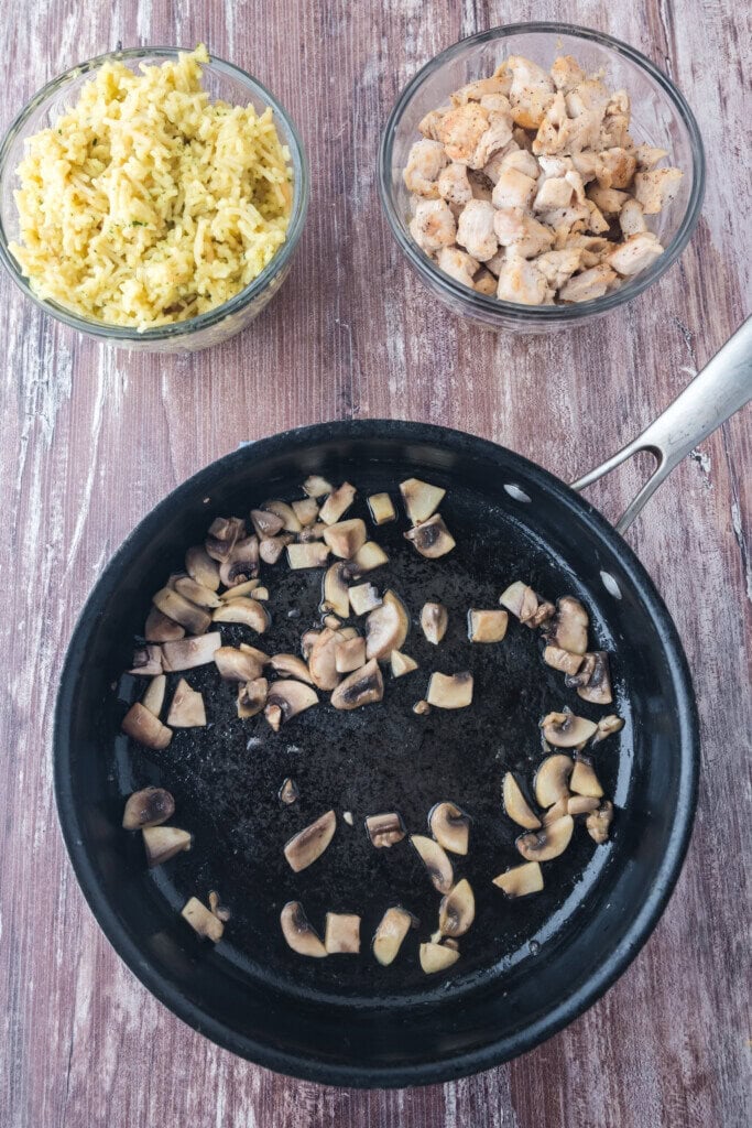 A black skillet with sautรฉed mushrooms sits on a wooden surface. Nearby are two glass bowls: one with yellow rice and the other with cooked chicken pieces.