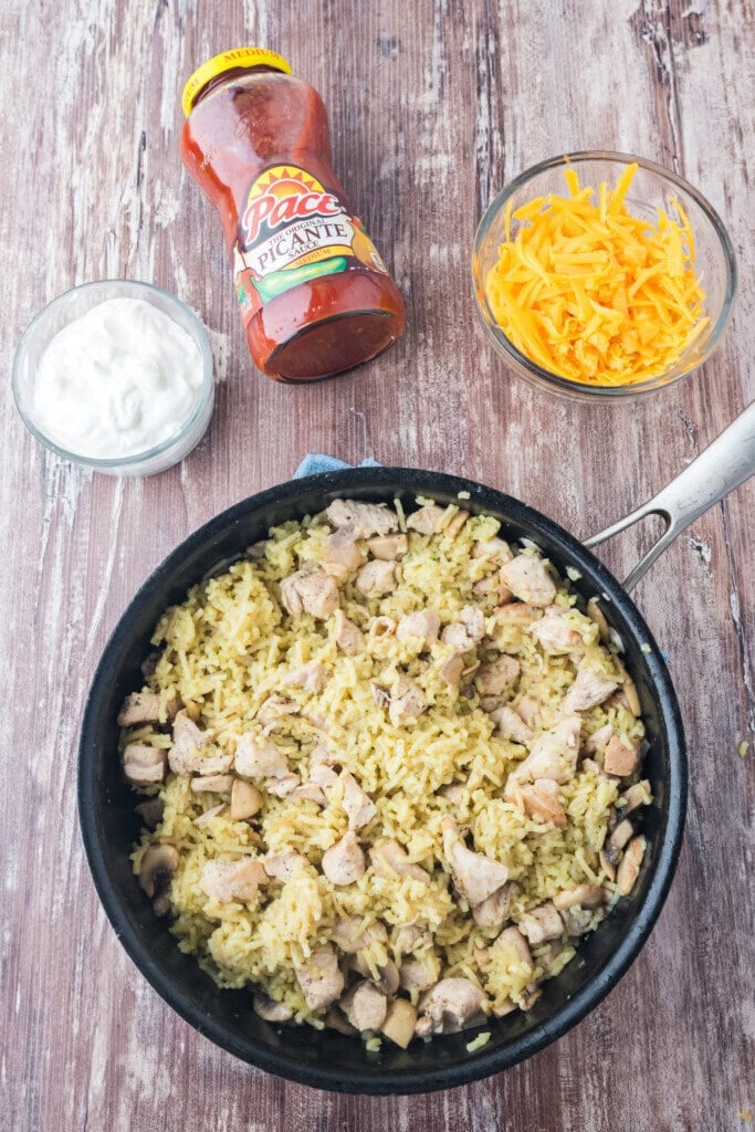 A skillet filled with chicken and rice sits on a wooden table, next to small bowls of shredded cheddar cheese and sour cream, and a jar of Pace Picante salsa.