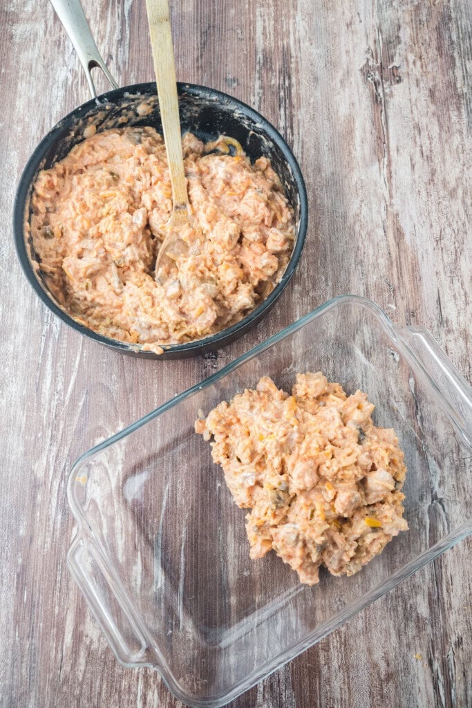 A pan of creamy, mixed casserole filling sits on a wooden surface with a wooden spoon inside. Some of the mixture has been transferred into a clear glass baking dish nearby.