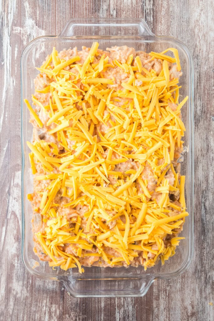A glass baking dish filled with a casserole mixture topped with shredded cheddar cheese, ready to be baked, sits on a rustic wooden surface.
