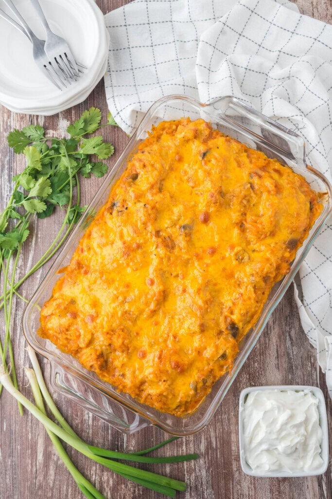 A glass baking dish filled with a cheesy, golden-brown casserole sits on a wooden table, surrounded by green onions, cilantro, a white napkin, plates with forks, and a small bowl of sour cream.