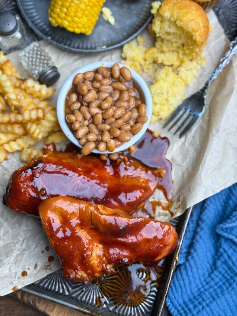 Barbecue chicken breasts with baked beans, crinkle-cut fries, a piece of corn on the cob, and crumbled cornbread on a tray, next to a blue textured cloth.