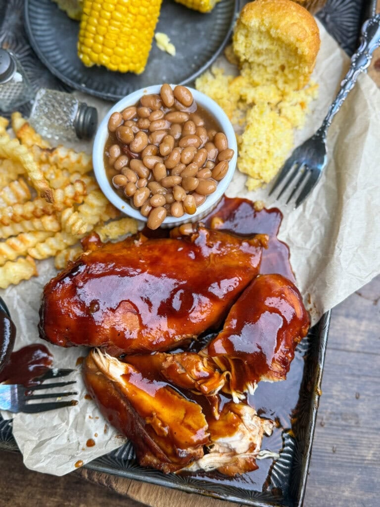 Barbecue chicken with sauce, crinkle fries, baked beans, cornbread, and corn on the cob served on a parchment-lined tray with forks and salt and pepper shakers nearby.