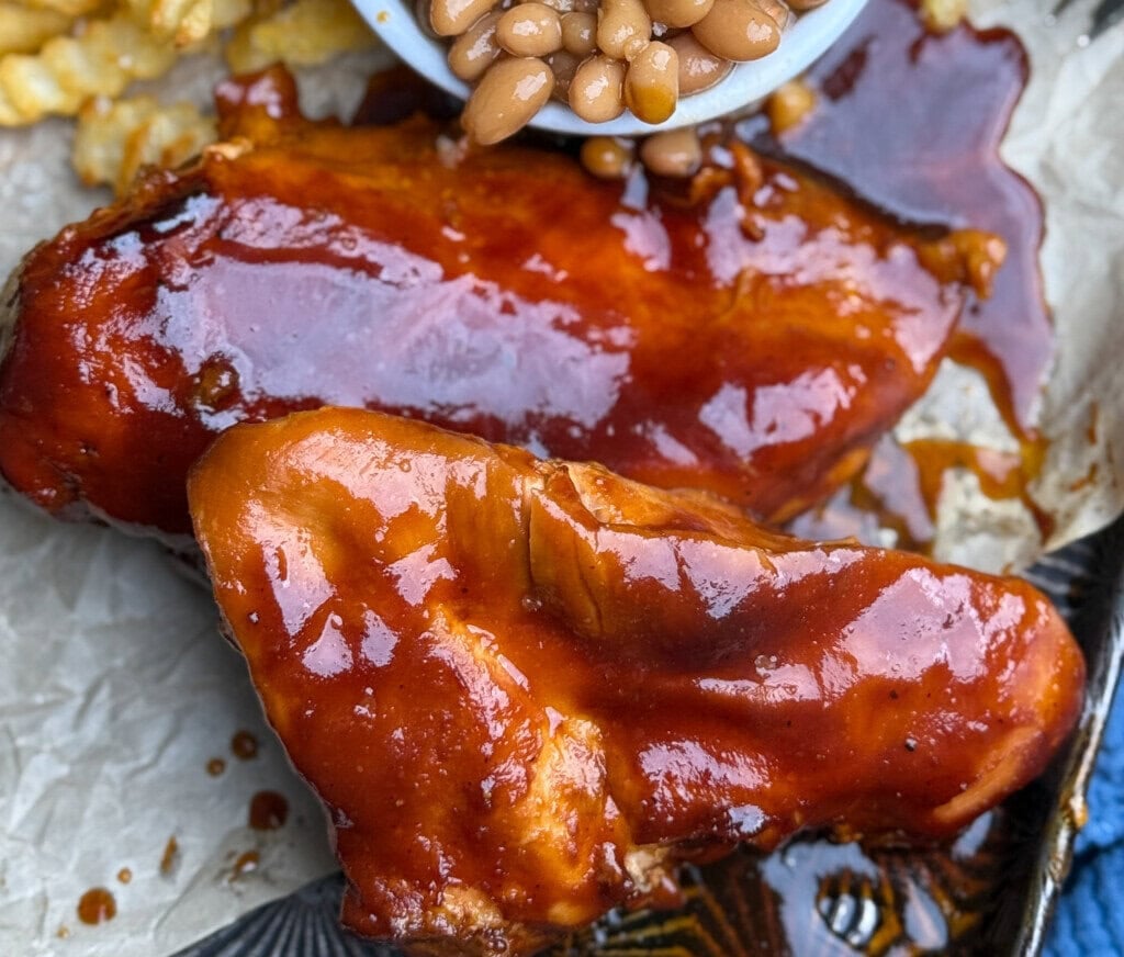 Two pieces of glazed barbecue chicken breast are served with crinkle-cut fries and baked beans on a parchment-lined tray, with sauce glistening on the chicken.