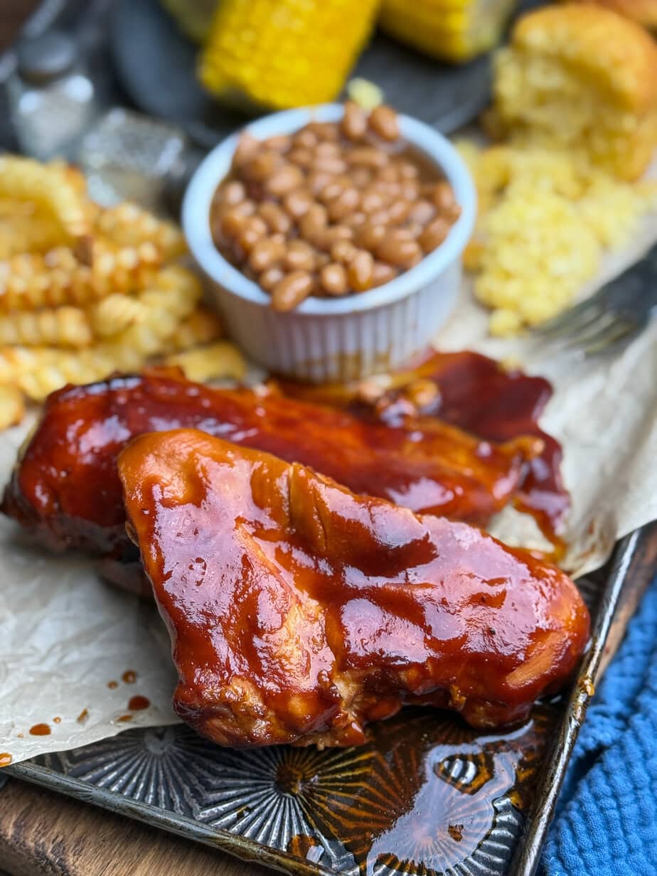 A close-up of barbecue chicken glazed with sauce, served with crinkle-cut fries, baked beans in a small bowl, corn on the cob, and cornbread on a metal tray.