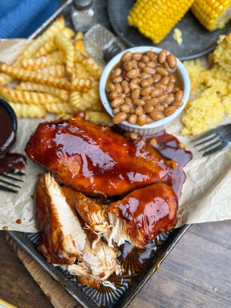 A plate with glazed barbecue chicken, crinkle-cut fries, baked beans in a small bowl, cornbread, and corn on the cob, all served on parchment paper.