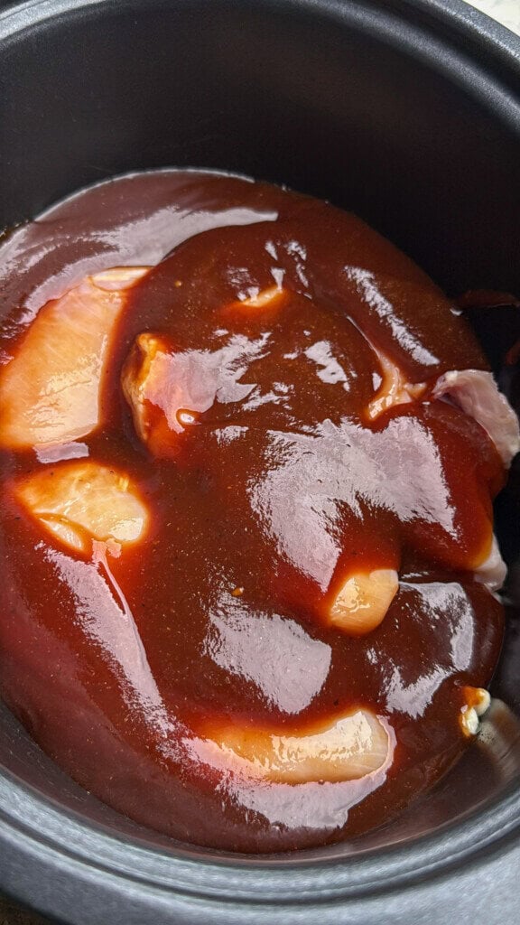 Raw chicken breasts are covered in a thick layer of dark barbecue sauce inside a black slow cooker.