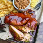 A plate with glazed barbecue chicken, crinkle-cut fries, baked beans in a small cup, and a piece of cornbread on parchment paper. A fork rests nearby, and the chicken is partially shredded.
