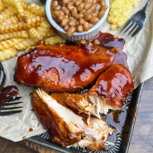 A plate with glazed barbecue chicken, crinkle-cut fries, baked beans in a small cup, and a piece of cornbread on parchment paper. A fork rests nearby, and the chicken is partially shredded.