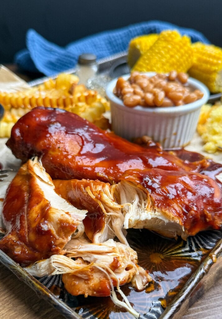 A plate with barbecue-glazed chicken, fries, baked beans in a ramekin, corn on the cob, and cornbread, set on a wooden table with a blue cloth in the background.
