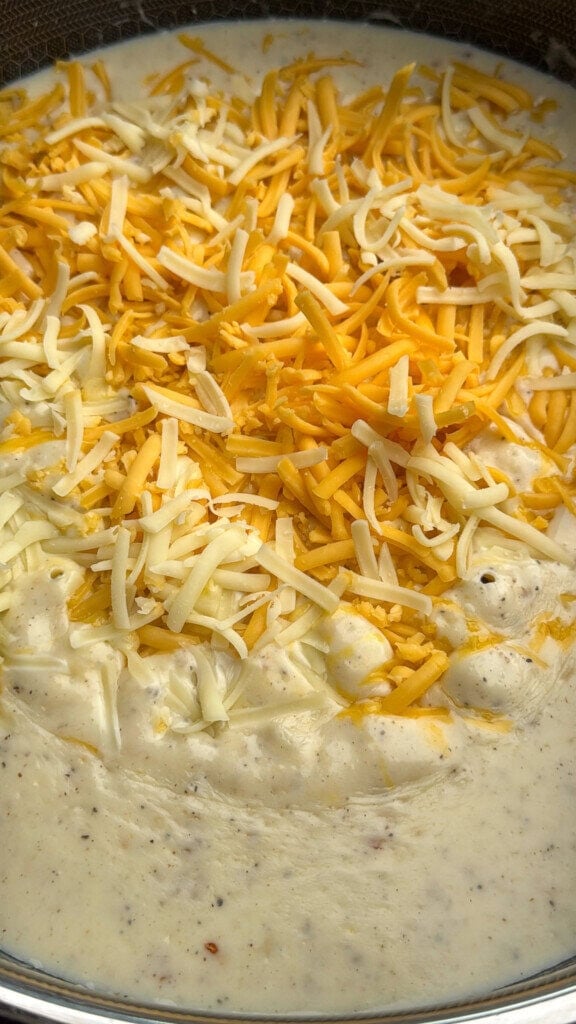 A close-up of a creamy, bubbling sauce topped with shredded cheddar and mozzarella cheese, about to be mixed in. The sauce appears thick and speckled with seasoning.
