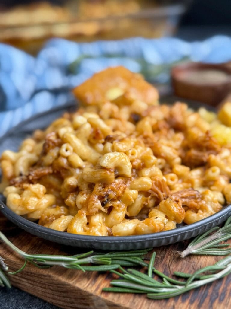 A close-up of a plate of macaroni and cheese mixed with pulled pork, served on a wooden board with fresh rosemary and a piece of cornbread in the background.