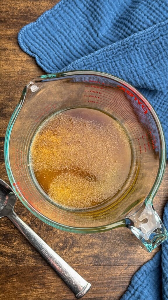A glass measuring cup filled with a vinegar and garlic powder. It sits on a wooden surface next to a silver spoon and a textured blue cloth.
