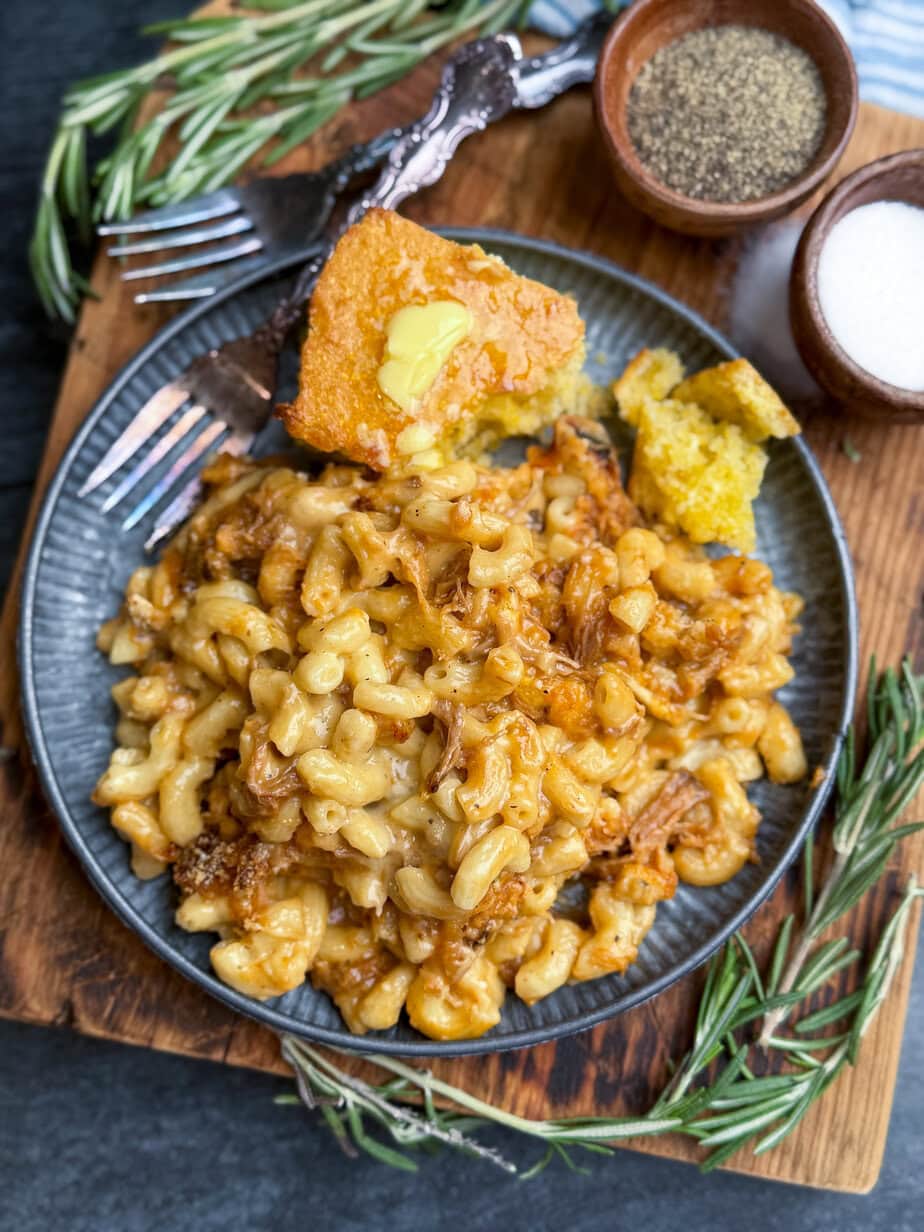 A plate of cheesy macaroni mixed with pulled pork, served with a slice of cornbread topped with melting butter. Fresh rosemary, a fork, and bowls of salt and pepper are nearby on a wooden board.