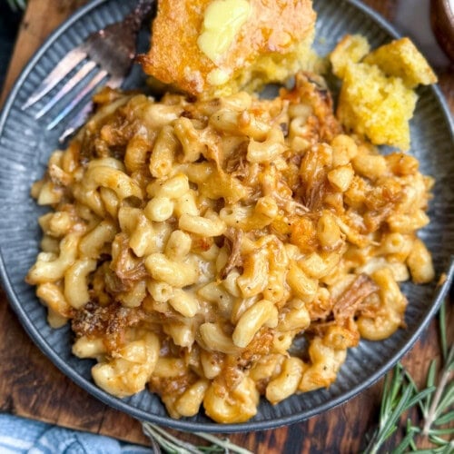 A plate of creamy macaroni and cheese mixed with pulled pork, served with a piece of buttered cornbread on the side. Fresh rosemary and a striped blue napkin are nearby.