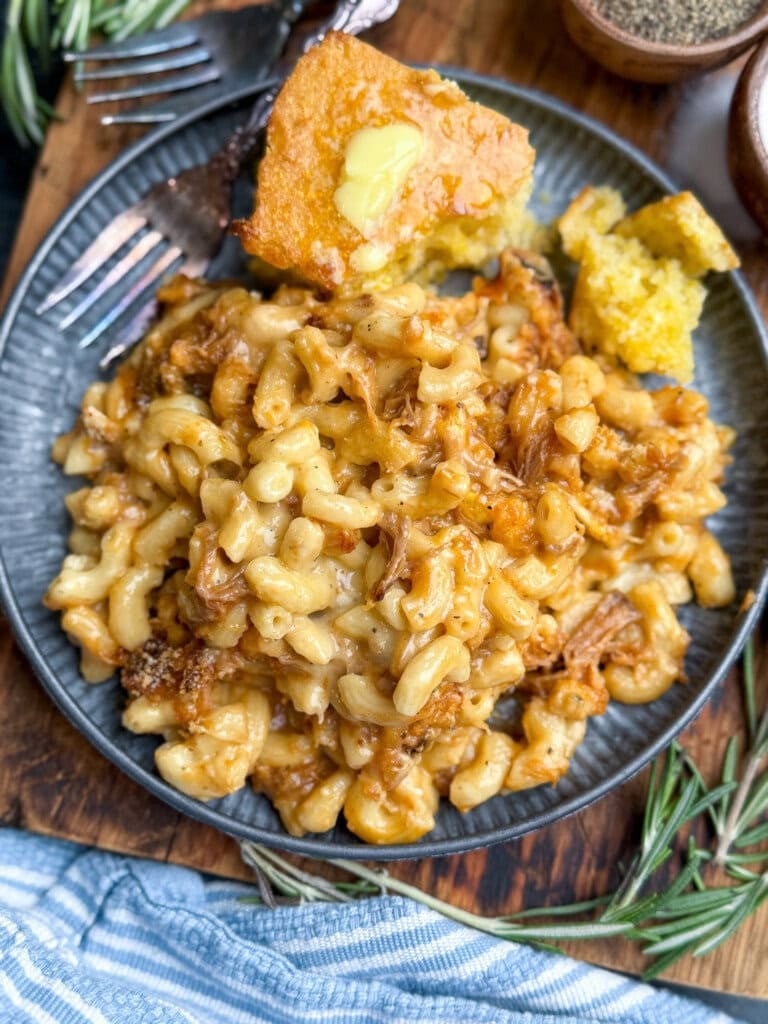 A plate of creamy macaroni and cheese mixed with pulled pork, served with a piece of buttered cornbread on the side. Fresh rosemary and a striped blue napkin are nearby.