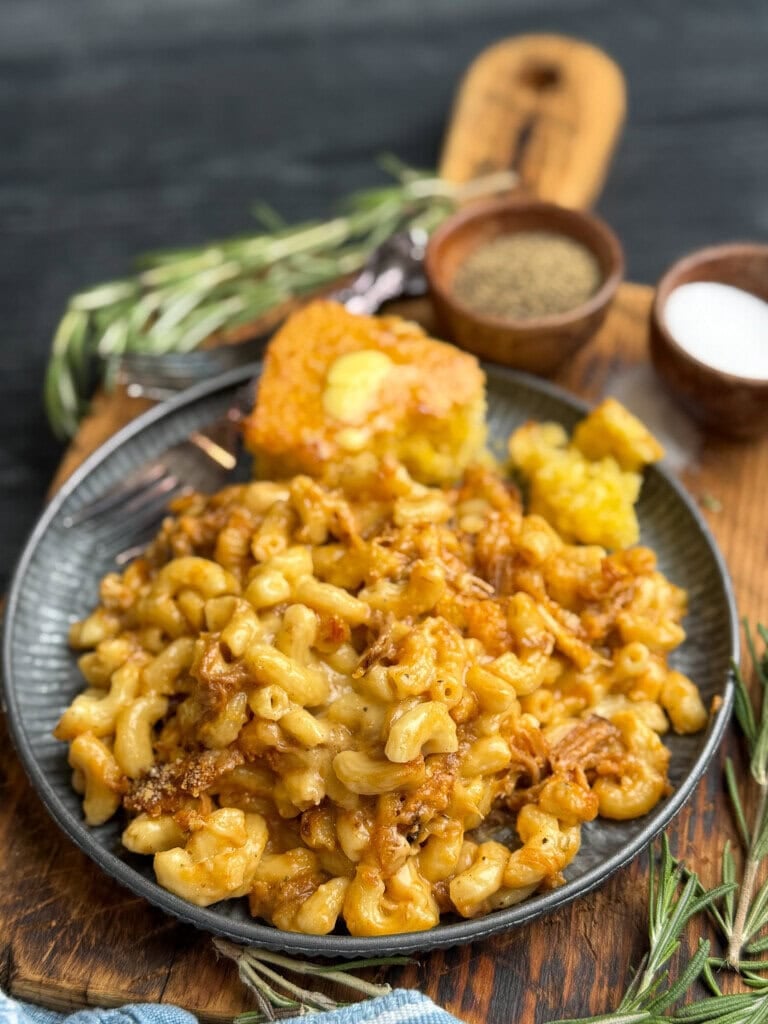 A plate of baked macaroni and cheese with shredded meat, served with a piece of cornbread on a wooden board. Small bowls of salt and pepper, sprigs of rosemary, and a blue cloth are nearby.