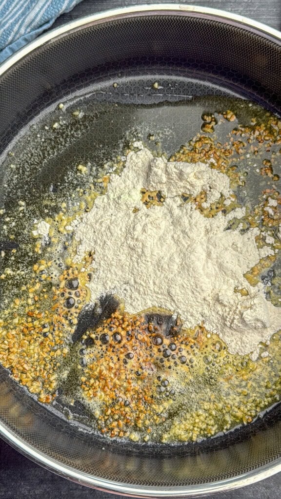 A close-up of a frying pan with sizzling oil, minced garlic, browned bits, and a mound of flour being cooked together.