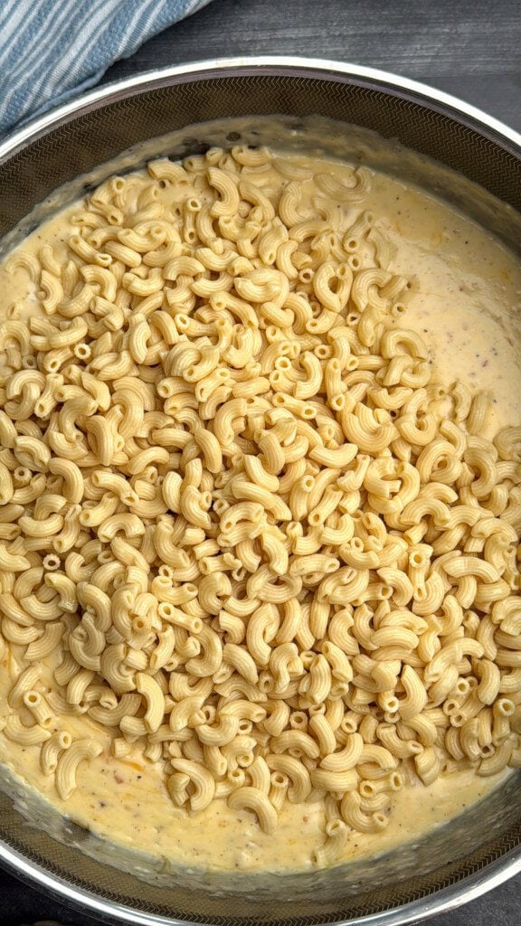 A close-up of cooked elbow macaroni in a creamy yellow cheese sauce, sitting in a large metal pot on a dark surface, with a blue-striped cloth visible in the corner.