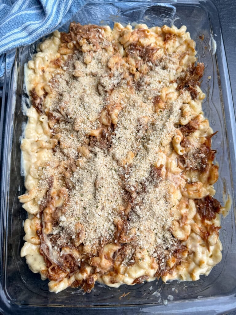 A glass baking dish filled with macaroni and cheese topped with shredded meat and sprinkled breadcrumbs, ready to be baked. A blue and white striped cloth is visible in the corner.