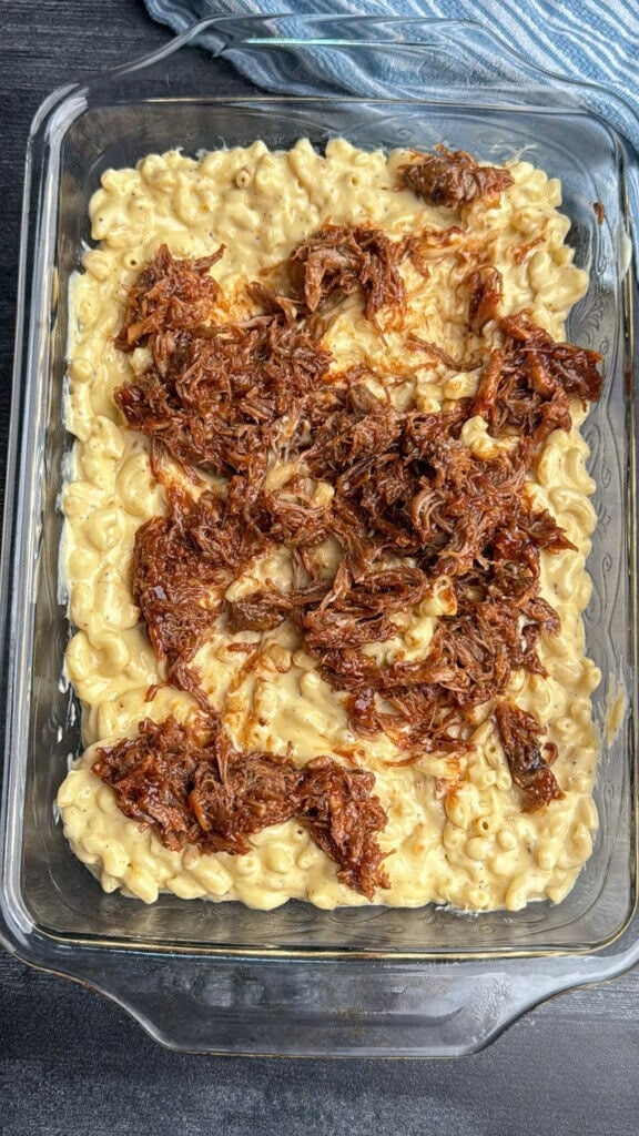 A glass baking dish filled with creamy macaroni and cheese, topped with shredded, saucy barbecue meat, sits on a dark surface. Part of a blue and white striped cloth is visible in the corner.