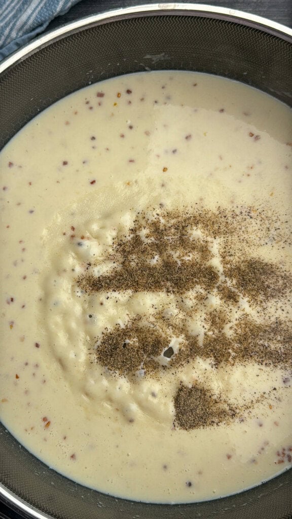A close-up of a creamy soup or sauce with visible pepper sprinkled on top, in a metal pot or bowl, with small bits throughout the mixture.