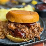 A pulled pork sandwich with barbecue sauce on a bun sits on a plate with baked beans and potato chips, with a small cup of barbecue sauce in the background.