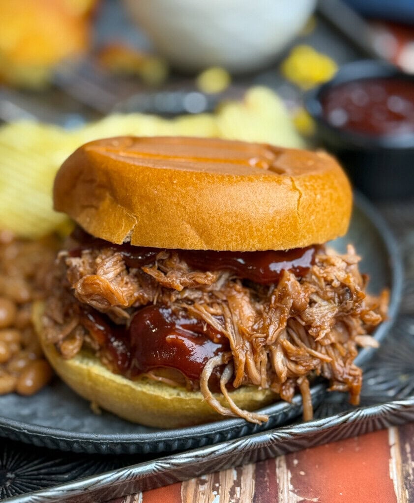 A pulled pork sandwich with barbecue sauce on a bun sits on a plate with baked beans and potato chips, with a small cup of barbecue sauce in the background.