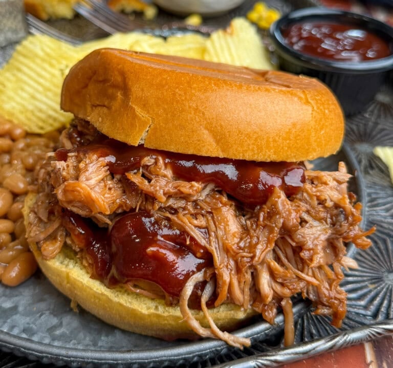 A pulled pork sandwich topped with barbecue sauce is served on a bun, accompanied by potato chips and baked beans on a plate, with a cup of ketchup in the background.