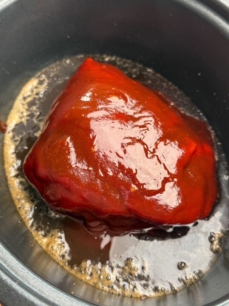 A piece of meat coated in a glossy bbq sauce sits in a slow cooker with dark root beer pooling around it.