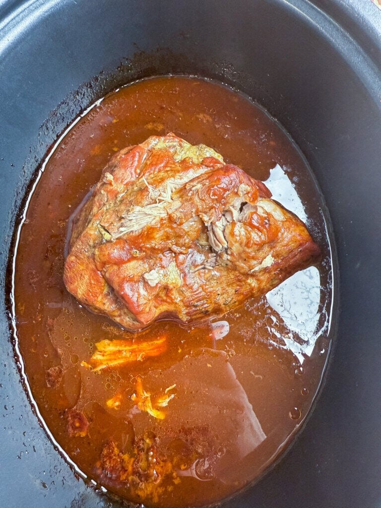 A large piece of cooked pork sits in a pool of reddish-brown sauce inside a slow cooker, making it perfect for a Crock Pot BBQ Pulled Pork Recipe With 3-Ingredients. Shredded pieces are visible in the flavorful sauce around the meat.