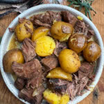 A bowl filled with tender chunks of sirloin steak and whole golden potatoes, garnished with herbs, sits on a wooden table next to sprigs of rosemary.