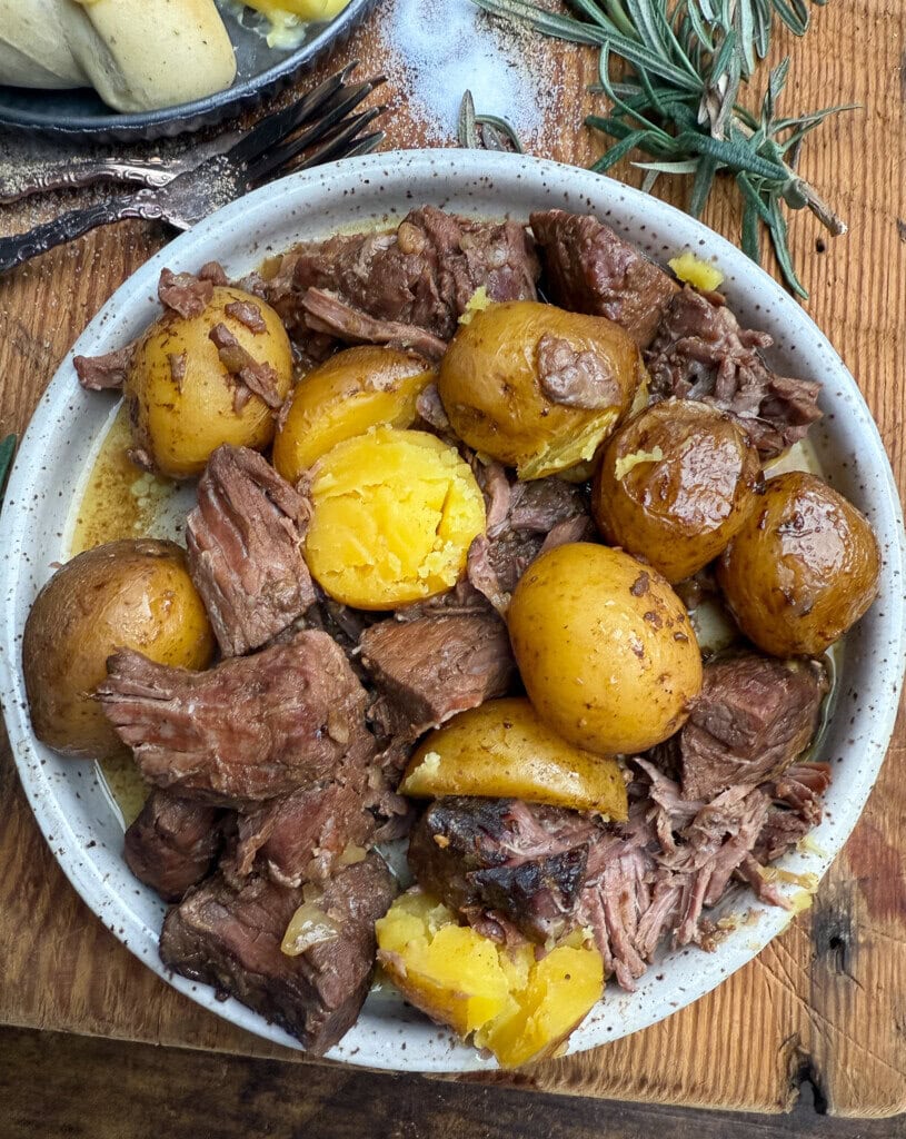 A bowl filled with tender chunks of pot roast and whole golden potatoes, garnished with herbs, sits on a wooden table next to sprigs of rosemary.