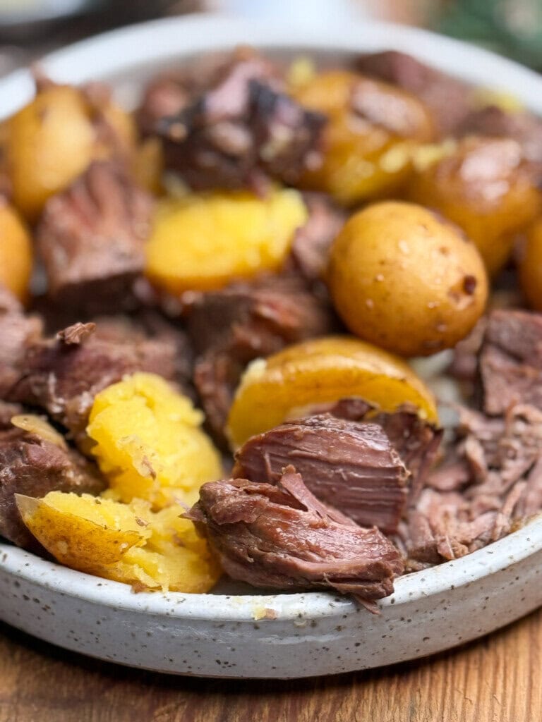 A close-up of a plate filled with tender chunks of beef and halved yellow potatoes, all cooked and slightly glistening, set on a wooden surface.