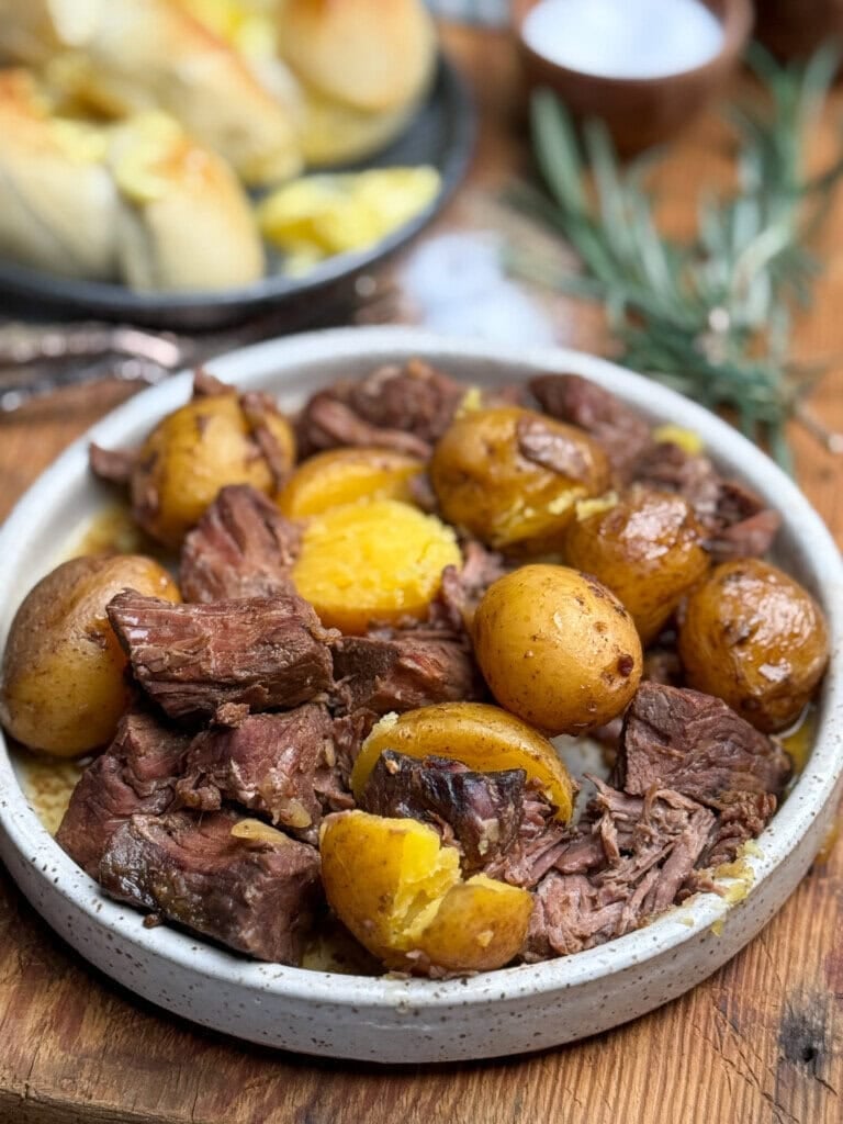 A ceramic plate filled with tender pieces of sirloin steak and whole golden potatoes, some potatoes split open, sitting on a wooden table with herbs and bread rolls in the background.