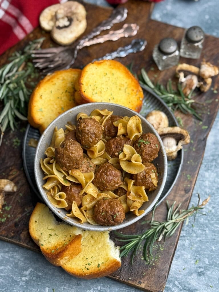 A bowl of pasta noodles and meatballs is surrounded by slices of garlic bread, fresh rosemary, mushrooms, and a set of forks on a wooden board. Salt and pepper shakers are in the background.