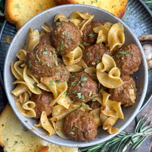 A bowl of egg noodles and meatballs garnished with herbs, served with slices of garlic bread and sprigs of fresh rosemary on the side.