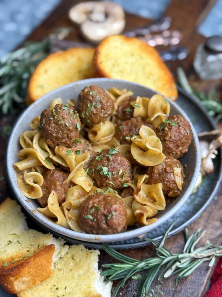 A bowl of egg noodles with meatballs, garnished with herbs, is served with slices of garlic bread. Rosemary sprigs and mushrooms are placed nearby on a wooden surface.