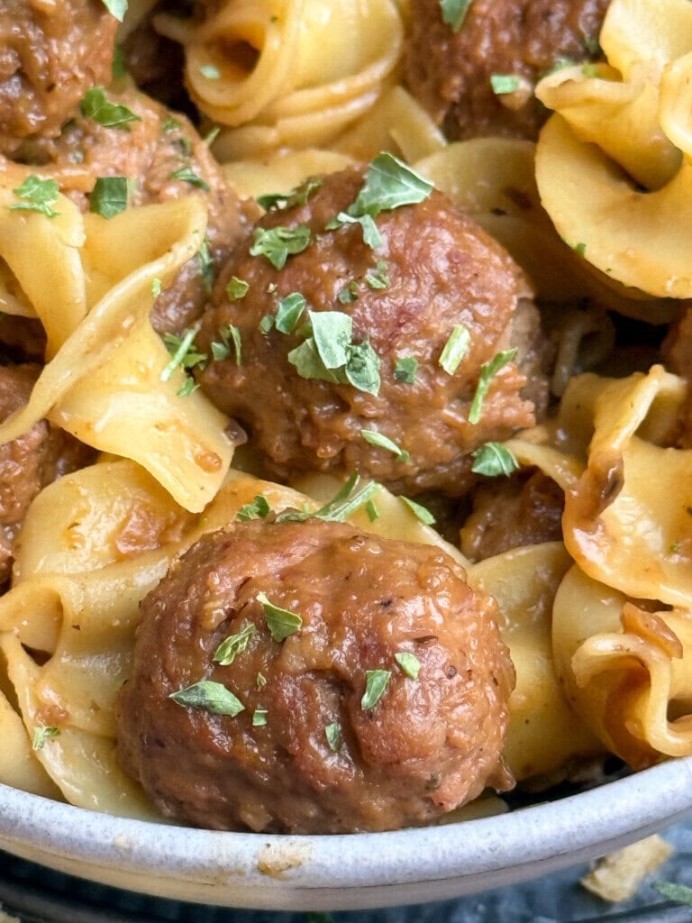 Close-up of cooked pasta noodles with browned meatballs, garnished with chopped green herbs. The dish appears saucy and flavorful, presented in a bowl.