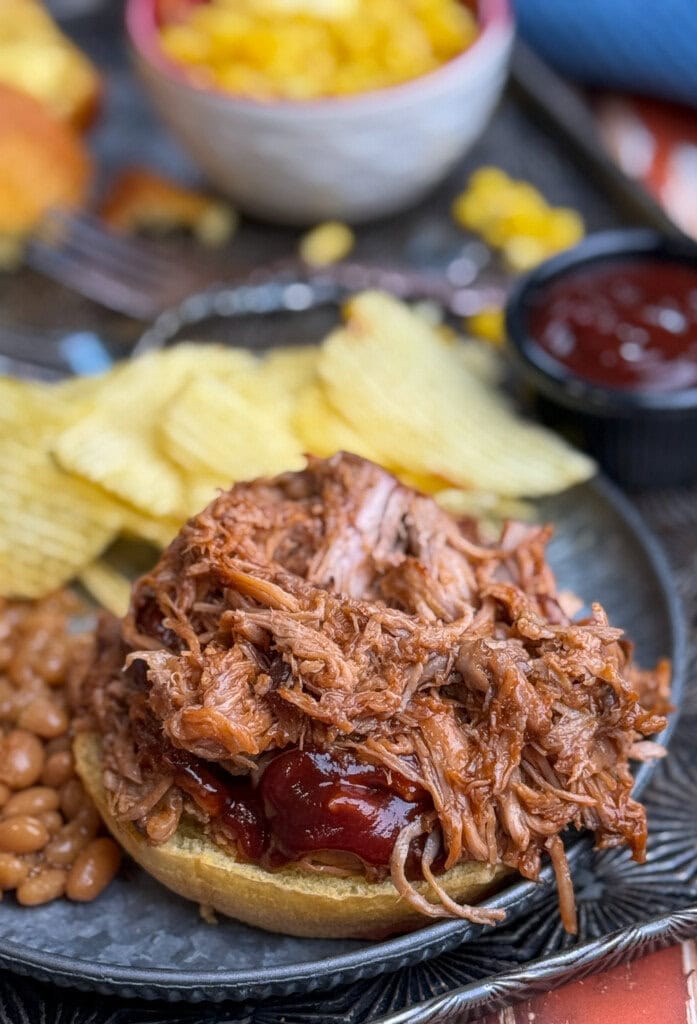A pulled pork sandwich with barbecue sauce on a bun, served on a plate with baked beans, potato chips, and a small cup of barbecue sauce. A bowl of corn is in the background.