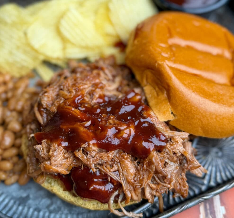 A pulled pork sandwich with barbecue sauce on a bun, served with baked beans and potato chips on a plate.