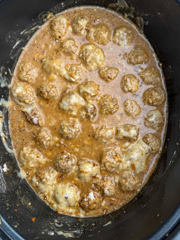 Meatballs simmering in a creamy brown sauce inside a slow cooker, with visible seasoning throughout the mixture.