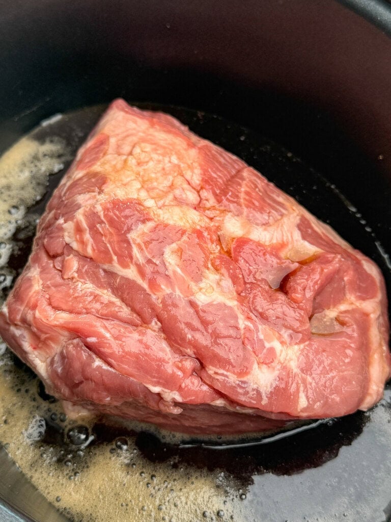 A raw piece of pork shoulder sits in a black crockpot with some root beer, ready for cooking.