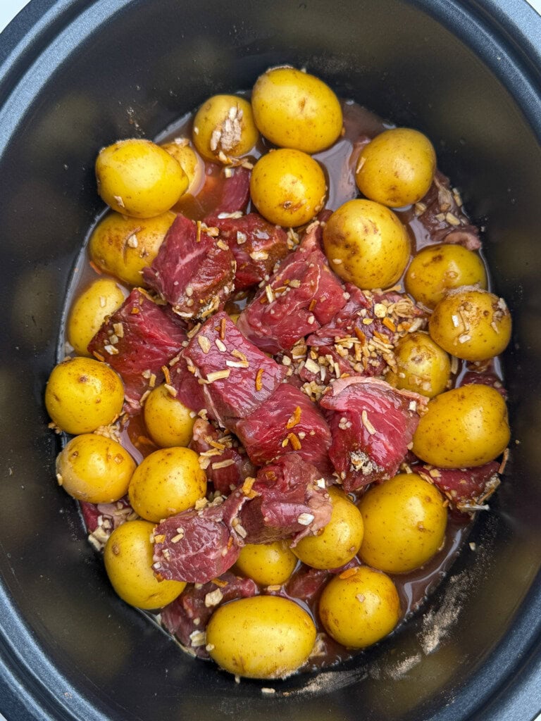 Raw beef chunks, whole baby potatoes, and minced onion sit in a slow cooker with broth, ready to be cooked. The ingredients are evenly distributed inside the black pot.