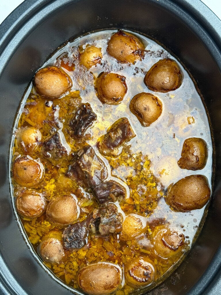 A slow cooker filled with whole baby potatoes, chunks of meat, and a rich broth, with visible herbs and seasonings.